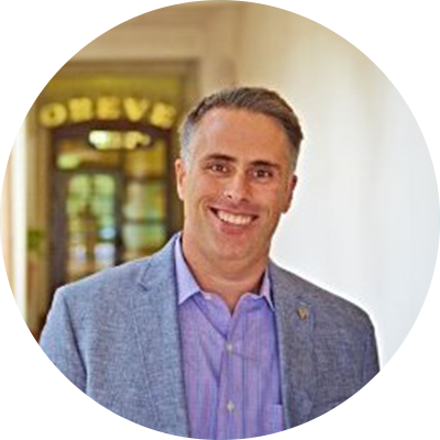 Smiling man in a blue suit jacket and purple shirt stands indoors with a blurred background.